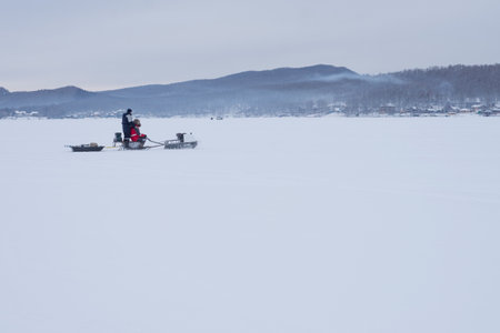 The snowmobile with fishermen is riding along the winter lake against the backdrop of the mountains.の写真素材