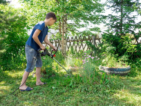 A boy, 11 years old, mows the lawn with an electric scythe near the trees in the yard of a house on a sunny summer day.の写真素材