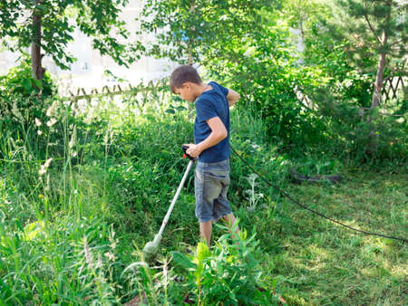 Boy, 11 years old, mows the lawn with an electric scythe near the trees in the yard of a house on a sunny summer day.の写真素材