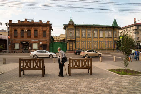 Krasnoyarsk, Krasnoyarsk Region, RF - July 19, 2021: A gray-haired man with a stick and a bag over his shoulder stands near the benches on the pavement opposite the old houses on Karl Marx Avenue.のeditorial素材