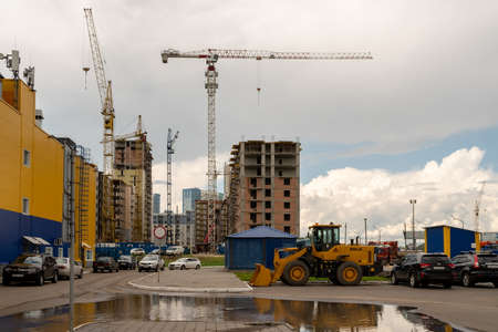 Krasnoyarsk, Krasnoyarsk Region, RF - July 20, 2021: Cars and a forklift are parked near the blue-yellow wall of a store against the backdrop of a construction site with tower cranes on a cloudy day.のeditorial素材