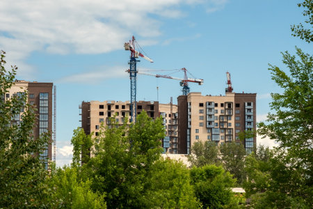 Krasnoyarsk, Krasnoyarsk Region, RF - July 1, 2021: Tower cranes in the construction of high-rise residential buildings are visible behind the trees on a sunny summer day.のeditorial素材