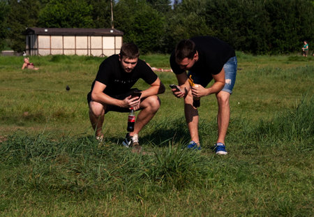 Krasnoyarsk, Krasnoyarsk Region, RF - July 24, 2021: Two young men take pictures of a wild gopher on a cell phone in a meadow with people resting among the green grass in summer.のeditorial素材