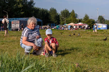 Krasnoyarsk, Krasnoyarsk Region, RF - July 24, 2021: A boy, sitting next to his grandmother, holds out food to the gopher, in a meadow with pigeons against the backdrop of pavilions.のeditorial素材