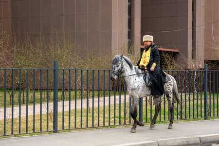 Krasnoyarsk, Krasnoyarsk Region, RF - May 9, 2021: A clergyman in a cassock and hat, with a cross on his chest, rides a horse along the sidewalk along a metal fence in the spring.のeditorial素材