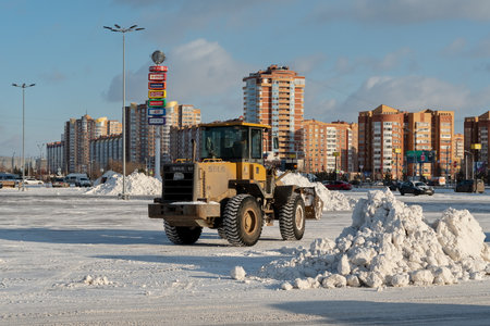 Krasnoyarsk, Krasnoyarsk Territory, RF - November 26, 2021: A bucket loader removes snow from snowdrifts in a city square after a snowfall against the backdrop of residential buildings on a sunny day.のeditorial素材