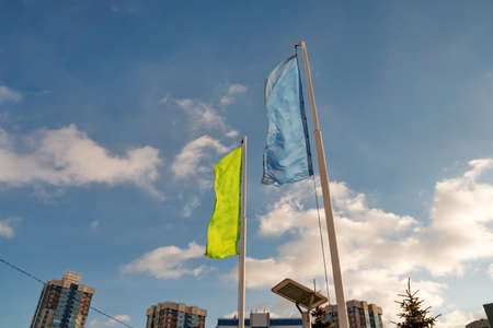 Two flags, yellow and blue, flutter in the wind over residential buildings across the sky on a sunny day.の写真素材