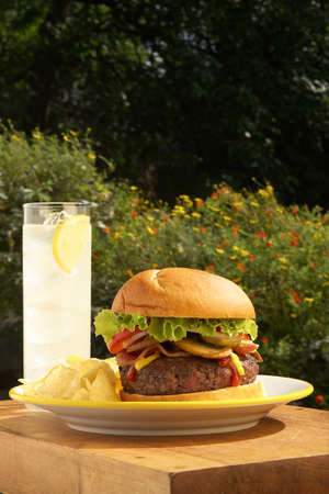 A close-up of lemonaid, potato chips and a hamburger topped with melted cheese, bacon, onion, tomato, lettuce, ketchup and mustard on wooden tabletop outdoors with flowers in the background and copy spaceの写真素材