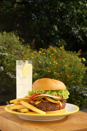 A close shot of lemonaid, french fries, and a hamburger topped mushrooms, bacon, onion, tomato, lettuce, ketchup and mustard on wooden tabletop outdoors with flowers in the background and copy space aboveの写真素材