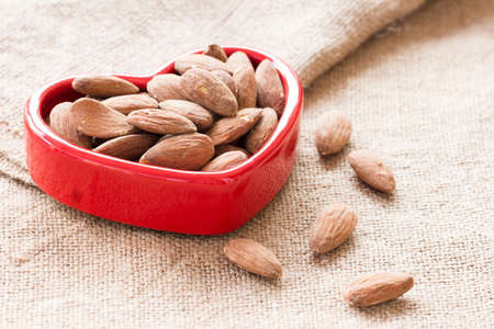 Almonds in red heart bowl  on sackcloth background, almonds in heart, brown almondsの写真素材