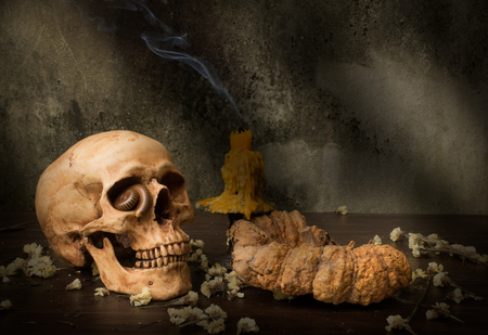 still life photography : human skull with worm in eye, candle have smoke, dry flowers and old pumpkin on old wall at back lighting from windowの写真素材