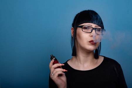 A beautiful girl with dark hair and glasses exquisitely smokes an electronic cigarette on a dark...