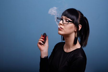 A beautiful girl with dark hair and glasses exquisitely smokes an electronic cigarette on a dark backgroundの写真素材