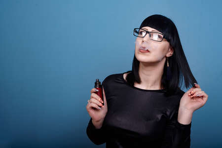 A beautiful girl with dark hair and glasses sensually smokes an electronic cigarette on a blue backgroundの写真素材