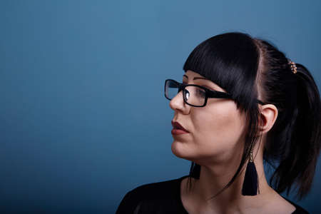 A beautiful girl with dark hair and glasses exquisitely smokes an electronic cigarette on a dark backgroundの写真素材