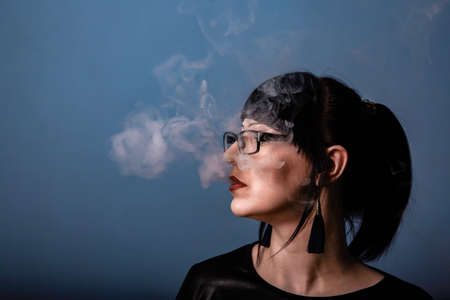 A beautiful girl with dark hair and glasses exquisitely smokes an electronic cigarette on a dark backgroundの写真素材