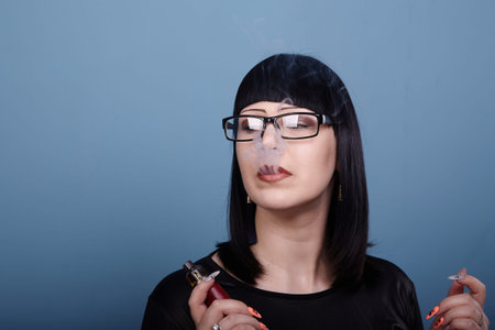 A beautiful girl with dark hair and glasses sensually smokes an electronic cigarette on a blue backgroundの写真素材