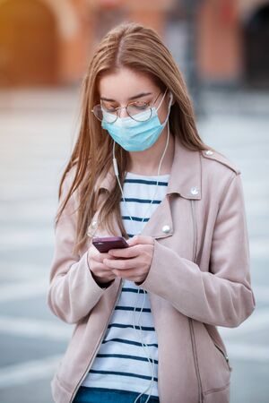 Young woman wearing medical face mask to protect herself from pollution, germs and coronavirus while texting or listen music in the cityの写真素材