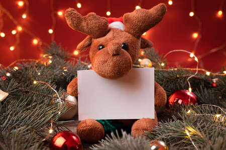 Image of a cute reindeer toy in santa hat holding a white,writeable card in a Christmas scenario.の写真素材
