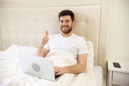 Man Working in Bed Showing Thumb Up. Man Working at Home. Man Using Laptop in Bed. Quarantine, Home Work, Medical Care.の写真素材