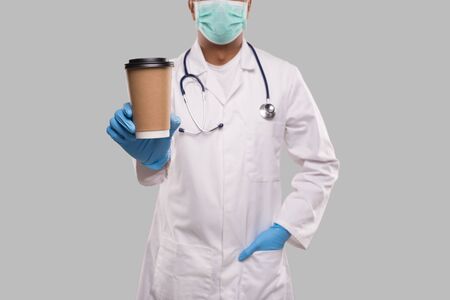 Indian Man Doctor Showing Coffee Take Away Cup Wearing Medical Mask and Gloves Close Up Isolated. Indian Doctor Holding Coffee To Go Cup. Medicの写真素材