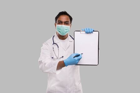 Doctor Pointing at Clipboard with Pen Wearing Medical Mask and Gloves Isolated. Indian Man Doctor Blank Clipboard in Handsの写真素材