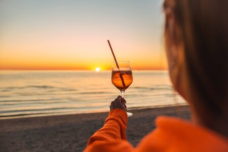 Girl Holding Glass of Drink on Beach at Beautyfull Romantic Sunset. Glass and Hand Close Up. View From Back. Girlの写真素材