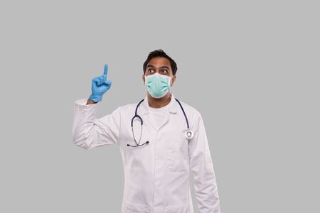 Indian Man Doctor Pointing Up and Watching Up Wearing Medical Mask and Gloves Isolatedの写真素材