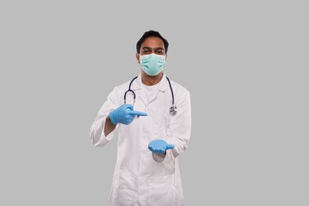 Doctor Pointing at Empty Open Hand in front of Chest Wearing Medical Mask and Gloves Isolated. Indian Man Doctor Advertisment Conceptの写真素材