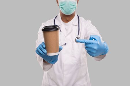 Indian Man Doctor Pointing at Coffee Take Away Cup Wearing Medical Mask and Gloves Close Up Isolated. Indian Doctor Holding Coffee To Go Cup. Medicの写真素材