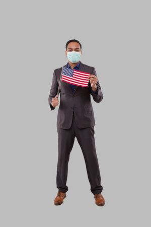 Business Man Holding American Flag Wearing Medical Mask. Indian Businessman Standing Full Length with USA Flag in Handsの写真素材