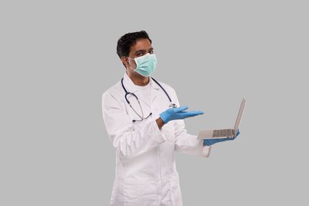 Doctor Pointing at Laptop Wearing Medical Mask and Gloves Isolated. Male Indian Doctor with Laptop in Hands.の写真素材