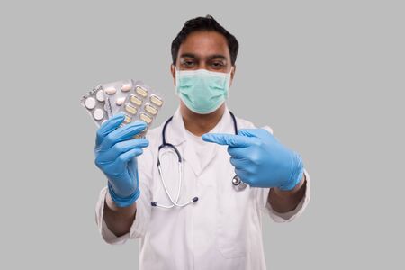 Man Doctor Pointing at Pills Wearing Medical Mask and Gloves. Doctor Holding Tablets. Indian Man Doctor Isolated.の写真素材
