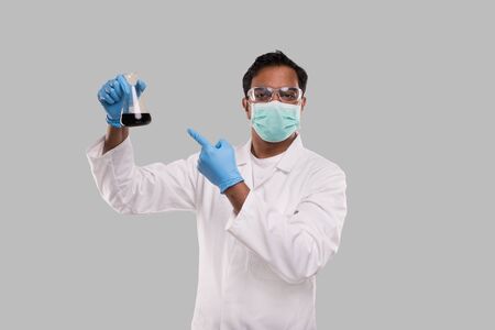 Male Doctor Pointing at Flask with Colorfull Liquid Wearing Medical Mask, Gloves and Glasses Isolated. Science, Medical, Virus Concept. Indian Man Doctor Smilling.の写真素材