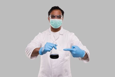 Male Doctor Pointing at Flask with Colorfull Liquid Wearing Medical Mask, Gloves and Glasses Isolated. Science, Medical, Virus Concept. Indian Man Doctor Smilling.の写真素材
