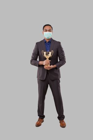 Businessman Holding Trophy Wearing Medical Mask. Indian Businessman Standing Full Length with Trophy in Handsの写真素材