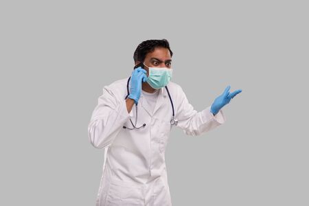 Doctor Angry Talking on Phone Wearing Medical Mask and Gloves. Indian Man Doctor with Phone Isolatedの写真素材