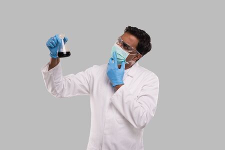 Male Doctor Examing Flask with Colorfull Liquid Wearing Medical Mask, Gloves and Glasses Isolated. Science, Medical, Virus Concept. Indian Man Doctor Smilling.の写真素材