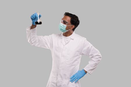 Male Doctor Examing Flask with Colorfull Liquid Wearing Medical Mask, Gloves and Glasses Isolated. Science, Medical, Virus Concept. Indian Man Doctor Smilling.の写真素材