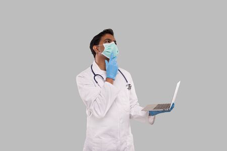 Doctor Using Laptop Wearing Medical Mask and Gloves Thinking Watching up Isolated. Male Indian Doctor with Laptop in Hands.の写真素材