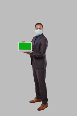 Businessman Showing Laptop Green Screen Isolated Wearing Medical Mask and Gloves. Indian Business man with Laptop in Hands. Online Business Conceptの写真素材