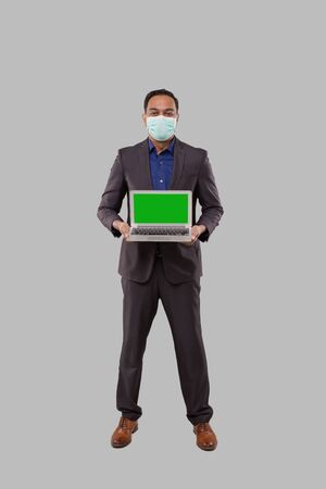 Businessman Showing Laptop Green Screen Isolated Wearing Medical Mask and Gloves. Indian Business man with Laptop in Hands. Online Business Conceptの写真素材