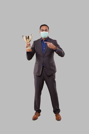 Businessman Pointing at Trophy Wearing Medical Mask. Indian Business man Standing Full Length with Trophy in Handsの写真素材
