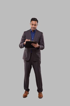 Businessman Writing in Clipboard Isolated. Indian Business man Standing Full Length with Clipboard in Handsの写真素材