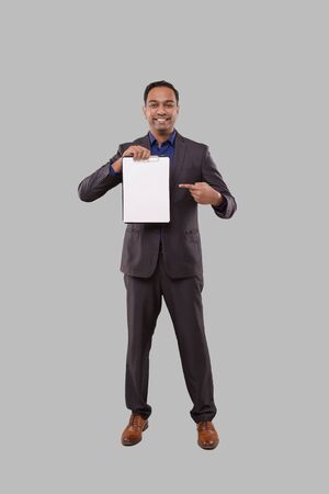 Businessman Pointing at Clipboard Watching in Camera Isolated. Indian Business man Standing Full Length with Clipboard in Handsの写真素材