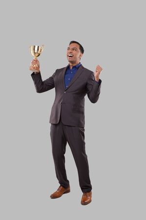 Businessman Very Happy and Excited, Raising arms, Celebrating a Victory or Success Holding Trophy. Winner Sign. Indian Business man Isolated with Trophy in Handsの写真素材