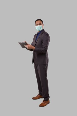 Businessman Using Tablet Wearing Medical Mask Isolated. Indian Business man Standing Full Length with Tablet in Handsの写真素材