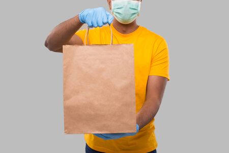 Delivery Man With Paper Bag in Hands Wearing Medical Mask and Gloves Isolated Close Up. Yellow Uniform Indian Delivery Boy. Home Food Delivery. Paper Bag Deliveryの写真素材