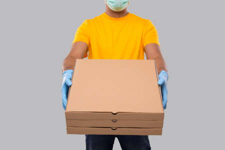 Delivery Man Three Pizza Box in Hands Wearing Medical Mask And Gloves Close Up Isolated. Yellow Tshirt Indian Delivery Boy. Man With Pizza in Hands. Deliveryの写真素材