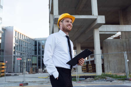 Businessman wearing Hardhat Holding Clipboard in hands. Builder Controlling House Building. Man Smilling near Construction Worksの写真素材